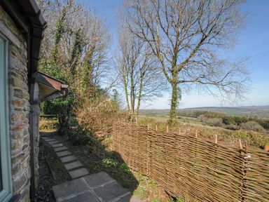 An outdoor path with stone tiles alongside a stone building next to a woven wooden fence and leafless trees at Rose Cottage in Callington