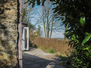 A stone wall with an open white door leading to a paved patio with a woven wooden fence and trees beyond at Rose Cottage in Callington