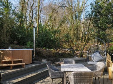 An outdoor wooden deck with a table and four chairs a hanging chair and a hot tub surrounded by trees at Rose Cottage in Callington
