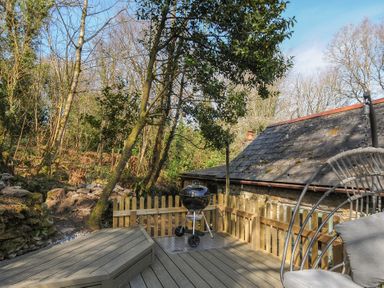 A wooden deck with a charcoal grill, wooden fence, and a cushioned hanging chair near trees and a stone building roof at Rose Cottage in Callington