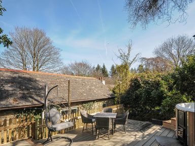 An outdoor deck with a hanging chair, table and chairs set, wooden fence, and hot tub surrounded by trees and a stone building at Rose Cottage in Callington