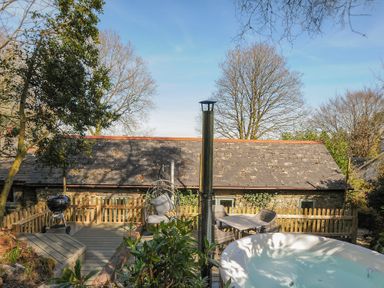 An outdoor deck with a hot tub table with chairs a hanging chair and a barbecue with trees and a stone building in the background at Rose Cottage in Callington