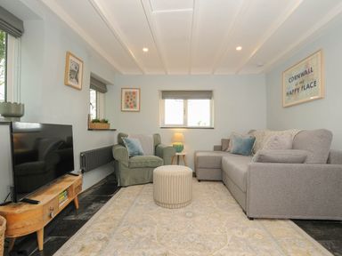 A living room with a grey sofa, green armchair, round ottoman, wooden TV stand, and framed art at Rose Cottage in Callington