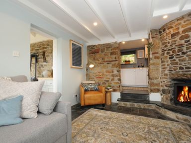 A living room with a gray sofa and leather chair near a stone fireplace and an open doorway to a kitchen at Rose Cottage in Callington