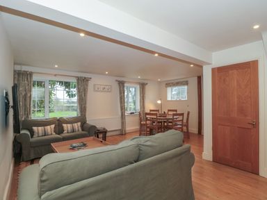 A living room with green sofas, wooden coffee table, dining table with chairs, windows with curtains, and a wooden door at Stable Cottage Lechlade-On-Thames