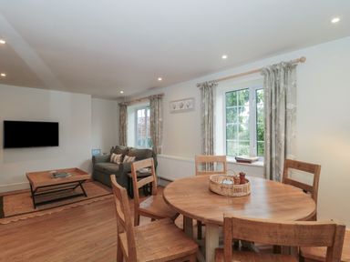 A living room with a wooden dining table and chairs a sofa TV and windows with curtains at Stable Cottage in Lechlade-On-Thames