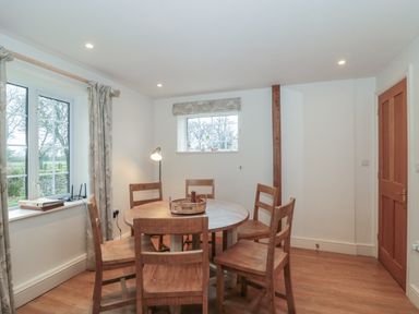 A dining room with a round wooden table and six wooden chairs near windows and a wooden door at Stable Cottage in Lechlade-On-Thames
