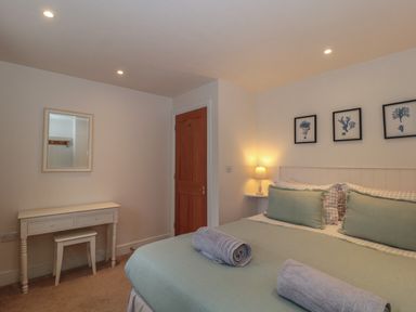 A bedroom with a bed with green pillows and rolled towels a wooden door a white desk with a stool a lamp and three framed artworks above the bed at Stable Cottage in Lechlade-On-Thames
