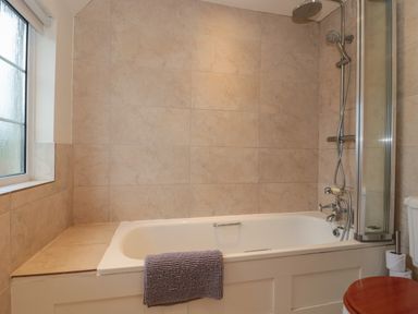 A bathroom with a bathtub a showerhead a window and a toilet with a wooden seat at Stable Cottage in Lechlade-On-Thames