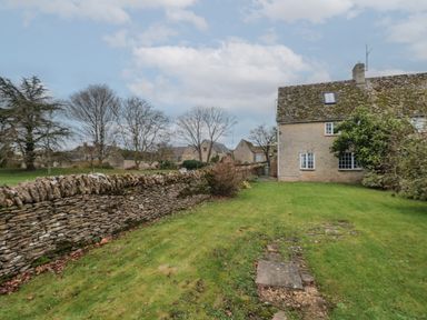 A stone house with a mossy roof next to a stone wall and a grassy yard with leafless trees at Stable Cottage in Lechlade-On-Thames