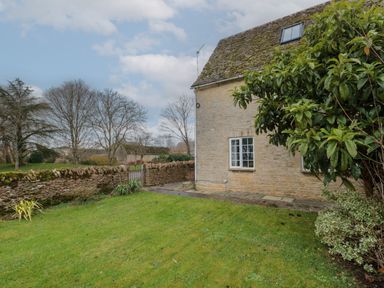 A garden with grass and trees next to a stone house and a stone wall with a gate at Stable Cottage in Lechlade-On-Thames