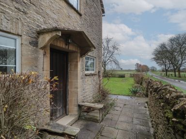 Stone house entrance with wooden door and small benches on a paved path next to a grassy area and a country road at Stable Cottage in Lechlade-On-Thames