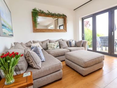 A living room with a gray sectional sofa and matching ottoman by glass doors with a wooden side table with red tulips and a large mirror above the sofa at Buzzards View in Dyserth
