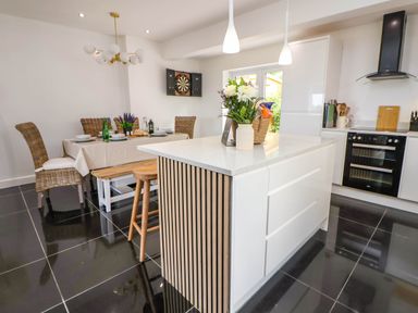 A kitchen with a white island and bar stools next to a dining table with wicker chairs and a dartboard on the wall at Buzzards View in Dyserth