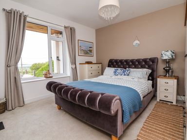 A bedroom with a tufted bed a window with curtains a chest of drawers and a nightstand with a lamp at Buzzards View in Dyserth