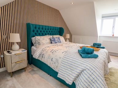 A bedroom with a teal upholstered bed and striped bedding two bedside tables with lamps and a window at Buzzards View in Dyserth