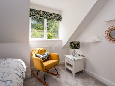 A bedroom corner with a yellow rocking chair next to a white bedside table with a lamp and a pillow at Buzzards View in Dyserth