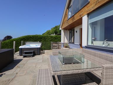 An outdoor patio with a wicker corner sofa set and glass table a hot tub and hedges at Buzzards View in Dyserth