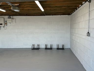 An empty room with white painted brick walls a gray floor ceiling with exposed wooden beams and three black metal bike racks attached to a wall at Buzzards View in Dyserth