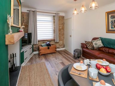 A living room with a dining table set with cups and eggs a brown leather sofa a television on a stand and a wooden chest by the window at Number Ten in Amble