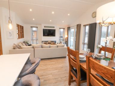 A living room with a large beige sofa a wall-mounted TV a dining table set with plates and wine glasses at 12A Lake Escape in Carnforth