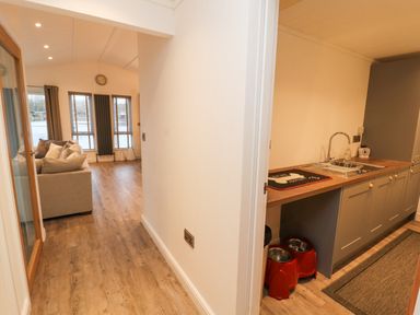 A hallway leading to a living room with a sofa and windows and a kitchen area with cabinets and a sink at 12A Lake Escape in Carnforth