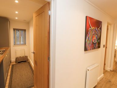 A hallway with a colorful painting of a cow on the wall and a partially open wooden door leading to a kitchen area with cabinets and pet food bowls at 12A Lake Escape in Carnforth