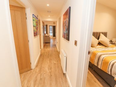 A hallway with wooden flooring and paintings on the walls leading to a living room with a sofa and a bedroom with a bed at 12A Lake Escape in Carnforth