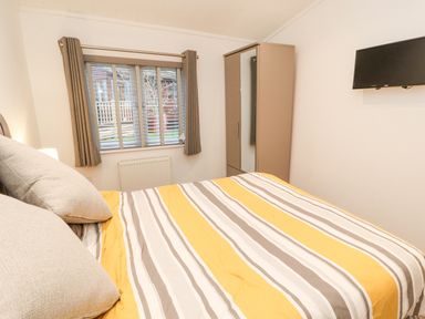 A bedroom with a bed covered in a striped yellow and gray bedspread a wardrobe a window with curtains and a wall mounted television at 12A Lake Escape in Carnforth