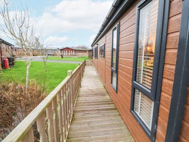 A wooden walkway with railing beside a brown building with windows overlooking grassy area and other buildings at 12A Lake Escape in Carnforth