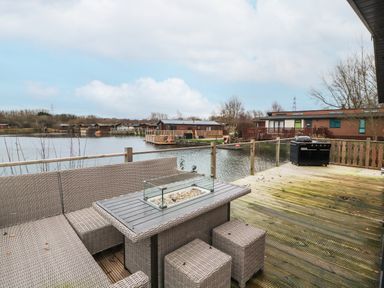 An outdoor deck with wicker furniture and a grill overlooking a lake with cabins at 12A Lake Escape in Carnforth