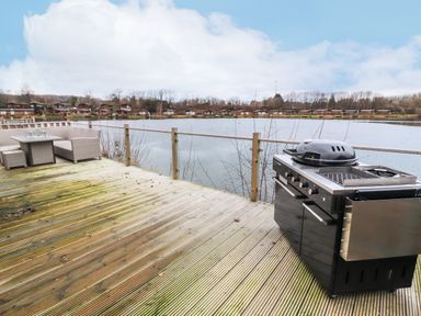 A wooden deck with a black barbecue grill and outdoor seating overlooking a lake with cabins across the water at 12A Lake Escape in Carnforth