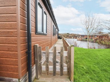 A wooden deck with a small gate next to a brown house by a lake with grass and leafless trees at 12A Lake Escape in Carnforth