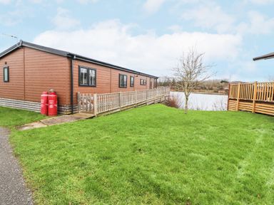 An exterior view of a brown mobile home with a wooden ramp next to a green lawn and a lake in the background at 12A Lake Escape in Carnforth