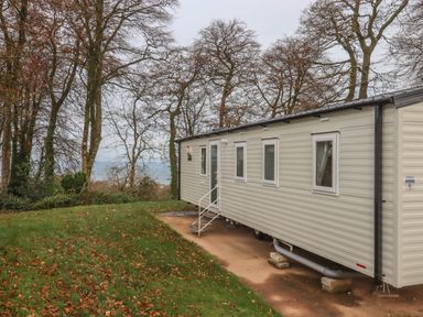 A mobile home on a grassy area with fallen leaves near trees and a body of water at 5 Manor Park in Bucks Cross Bideford