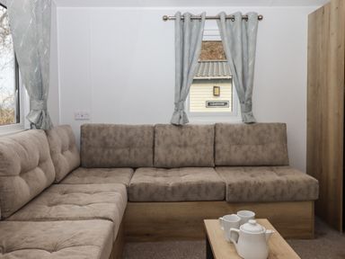 A living room with an L-shaped sofa a small coffee table with a teapot and cups and windows with curtains at 5 Manor Park in Bucks Cross Bideford