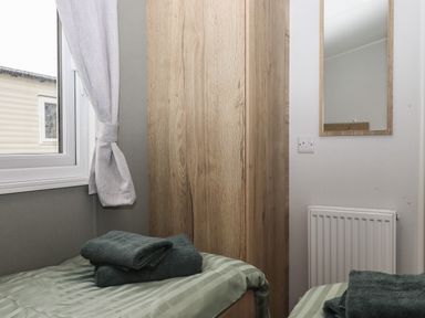 A bedroom with two beds folded towels a window curtain a wooden wardrobe a mirror on the wall and a radiator at 5 Manor Park in Bucks Cross Bideford