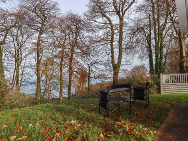 A grassy backyard with fallen leaves trees without leaves two black outdoor chairs and a wooden deck at 5 Manor Park Bucks Cross Bideford