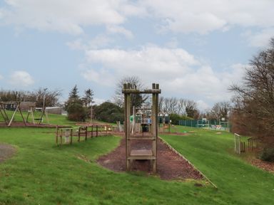 A playground with swings climbing structures and a fenced sports court in a grassy outdoor area at 5 Manor Park in Bucks Cross Bideford
