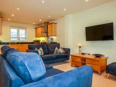 A living room with blue sofas a wooden coffee table with a lamp and a wall-mounted TV with a kitchen area in the background at Blue Horizon in Port Isaac