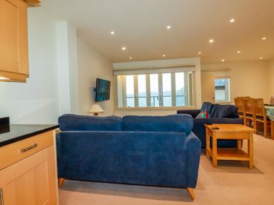 A living room with blue sofas a wooden side table a wall-mounted television and a dining area at Blue Horizon in Port Isaac
