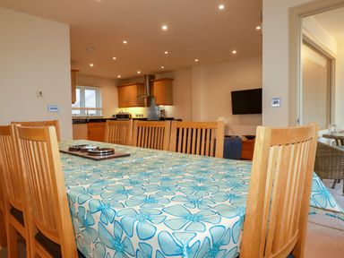 A dining area with wooden chairs and a table covered with a floral tablecloth adjacent to a kitchen with wooden cabinets and a wall-mounted TV at Blue Horizon in Port Isaac