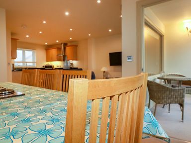 A dining area with a wooden table covered with a blue patterned tablecloth and wooden chairs next to a kitchen with wooden cabinets and a living area with a wall-mounted TV and a lamp at Blue Horizon in Port Isaac