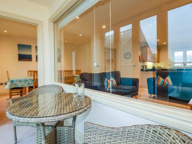 A seating area with a glass-top wicker table and two wicker chairs next to a large window showing a living room with two sofas and a dining area at Blue Horizon in Port Isaac