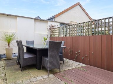 An outdoor dining area with a glass table and six wicker chairs on a paved patio with a planter and wooden fence at Blue Horizon in Port Isaac
