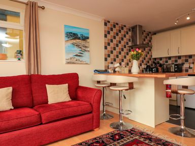 A living room with a red sofa and beige pillows next to a kitchen island with three white bar stools a tiled backsplash and a vase of flowers on the counter at Garden View in Hillhead near Brixham