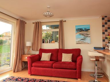 A living room with a red sofa two beige cushions a wooden side table with a lamp and a painting on the wall at Garden View Hillhead near Brixham