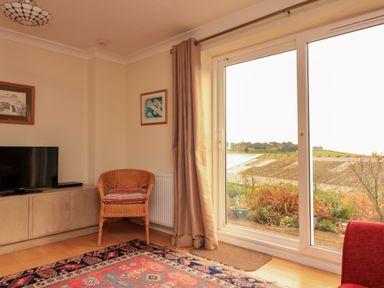 A corner of a living room with a wicker chair and cushion a television on a cabinet a sliding glass door with curtains and a view of a garden at Garden View in Hillhead near Brixham