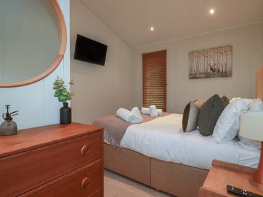 A bedroom with a bed pillows and towels a wooden dresser with a vase a round mirror a wall-mounted tv and a window with blinds at 3 Hedgerows Lanreath