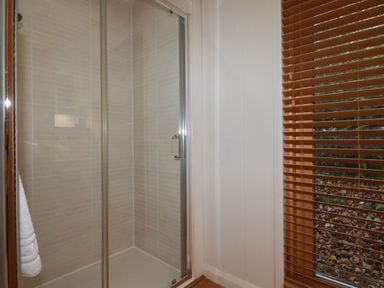 A bathroom shower with glass doors next to a window with wooden blinds at 3 Hedgerows in Lanreath